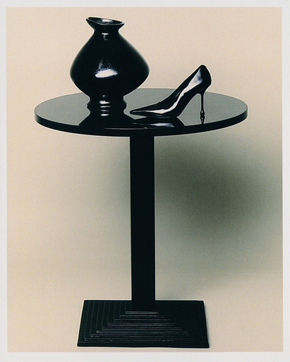 Black still life: a high-heeled shoe and vase on a sleek, round black table with a square base against a gradient cream backdrop.