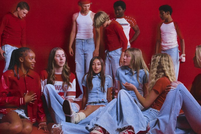 Diverse group of teenage models in jeans and red and white tops are posed together against a solid red backdrop in a studio setting.