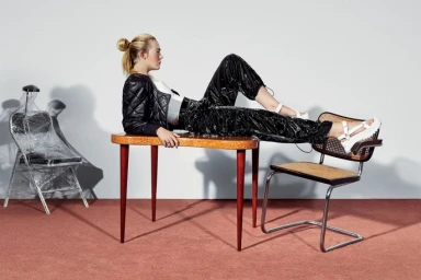 A blonde model styled by James Rene wears a black quilted jacket and shiny pants while reclining on a vintage wooden table.