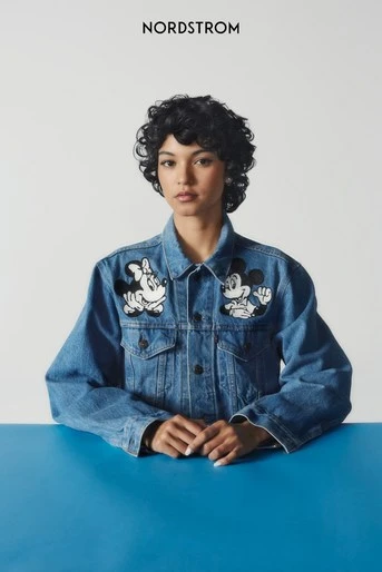 A young woman with curly hair wearing a denim jacket with cartoon character patches sits at a blue table in front of a white background.