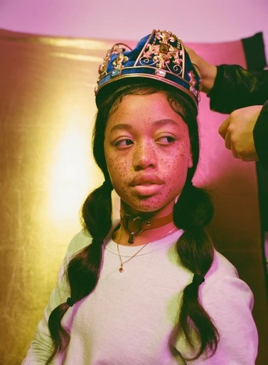 Portrait of a freckled young woman wearing an ornate blue crown. Hands are visible placing the crown. Pink and gold background.