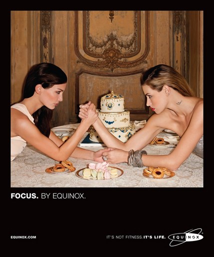 Two women arm wrestling at a table with desserts and an ornate cake in the background. This image is part of an Equinox campaign.