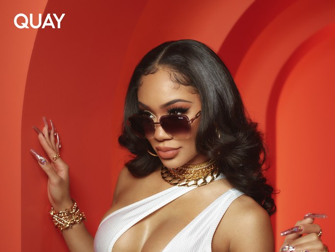 A stylish woman with long brown hair and sunglasses poses against a warm orange background, wearing gold jewelry and a white top.
