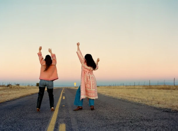 Two women with arms raised stand on a rural asphalt road at sunset wearing patterned coats and jeans.