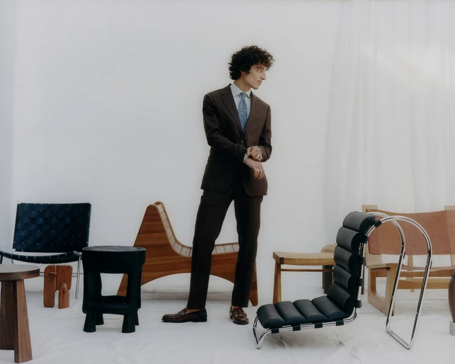A man in a brown suit stands near an arrangement of unique chairs in a light studio setting, captured in a medium shot.