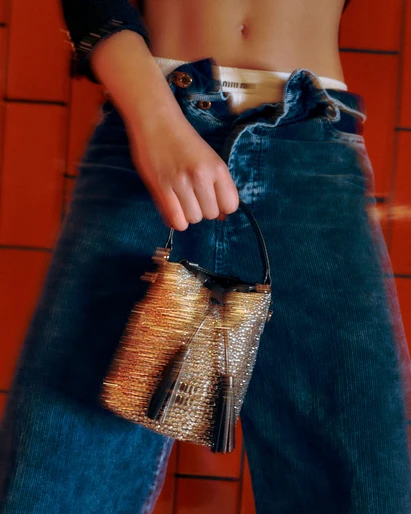 Woman holds a shiny gold purse, posing with unbuttoned dark blue jeans, visible white underwear band, and a red brick wall background.