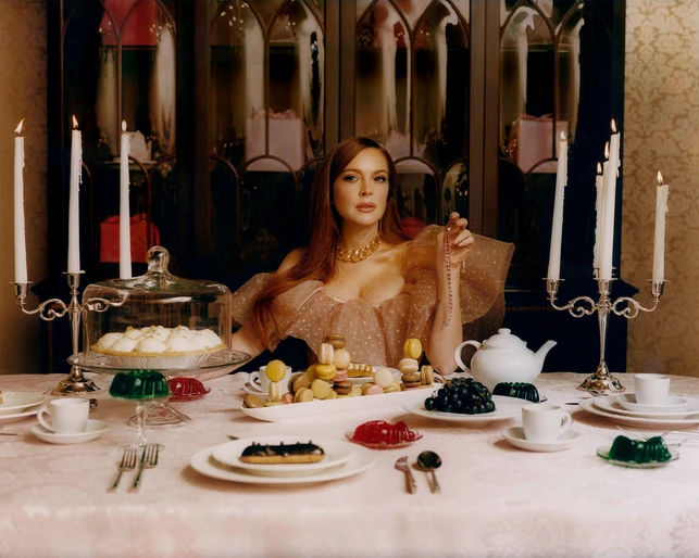 Adult woman at a formal table setting with desserts, gelatin, teapot, and candles, wearing an off-the-shoulder dress and jewelry.