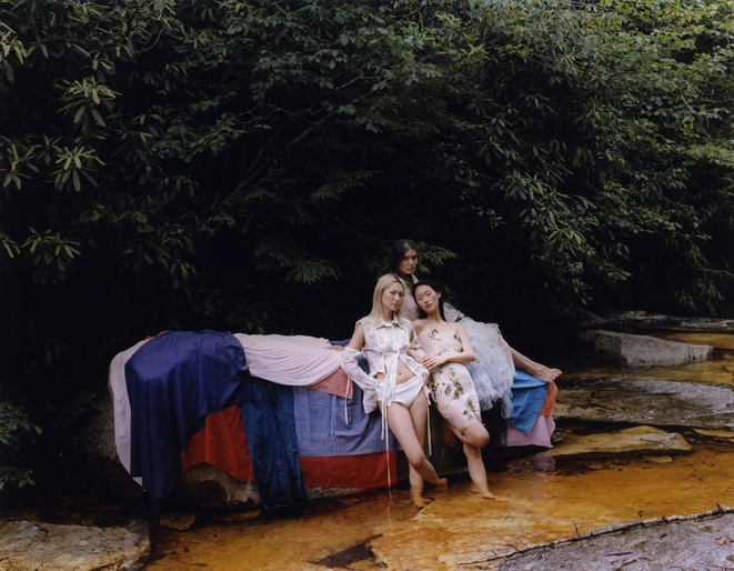 Three women pose on a makeshift bed of colorful fabrics outdoors surrounded by greenery. Fashion, summer aesthetic, and nature.