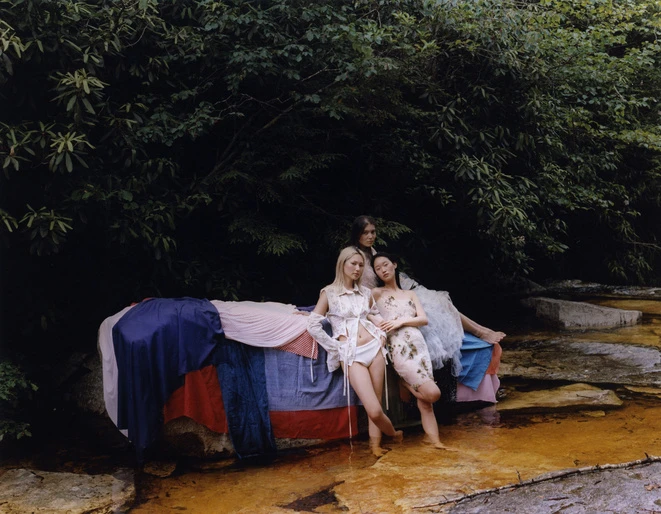 Three women pose on a makeshift bed of colorful fabrics outdoors surrounded by greenery. Fashion, summer aesthetic, and nature.
