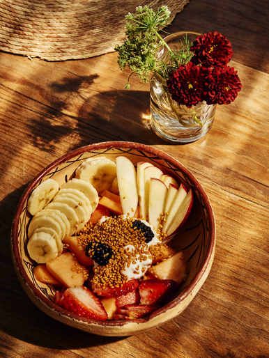 Overhead shot of a vibrant fruit bowl with strawberries, bananas, apples, papaya, and blackberry on a wooden table with flowers.