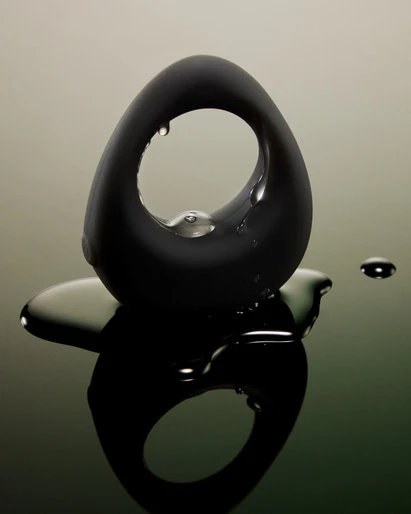 Sleek, black, ovoid object with a central hole, adorned with water droplets, reflecting on a glossy surface with a blurred background.