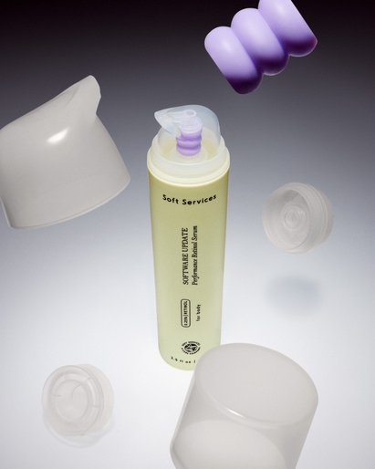 Still life shot of a light yellow Soft Services skincare bottle surrounded by floating white and purple lids on a gradient gray backdrop.