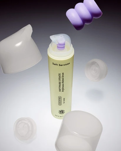 Still life shot of a light yellow Soft Services skincare bottle surrounded by floating white and purple lids on a gradient gray backdrop.