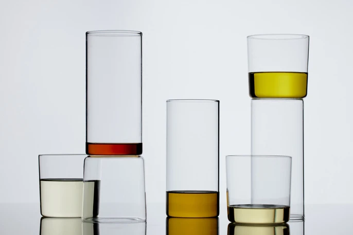 Still life of clear glasses with colorful liquids stacked in a minimalistic composition against a bright, neutral backdrop, studio shot.