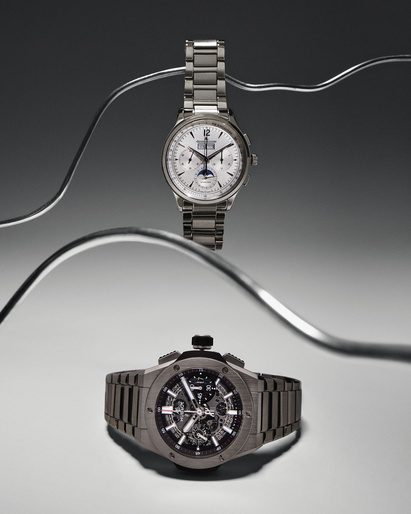 Two luxury wristwatches are displayed with curved metal rods on a light gray background, showcasing their detailed designs and metallic finishes.