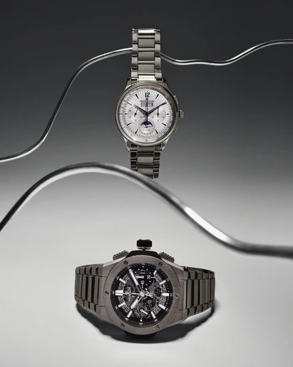 Two luxury wristwatches are displayed with curved metal rods on a light gray background, showcasing their detailed designs and metallic finishes.