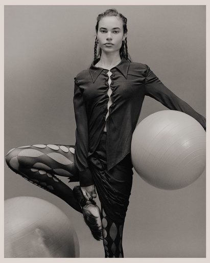 Monochrome studio shot of a female model in dark top and patterned tights, balancing with a large white exercise ball.