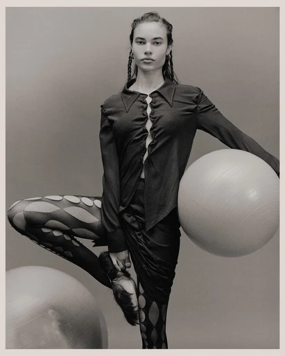 Monochrome studio shot of a female model in dark top and patterned tights, balancing with a large white exercise ball.