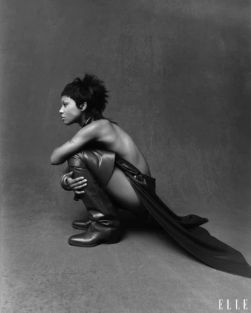 Monochrome profile portrait of a woman crouching wearing tall leather boots and a dark cape against a textured studio background.