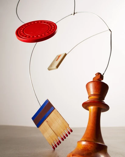 Conceptual mobile art with chess king, matchsticks, numbered tile, and red disc suspended by wires against a neutral backdrop.