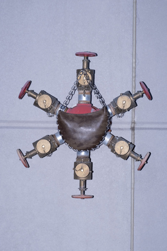 Brass industrial valve fixture with red accents hangs against a grey tiled wall, featuring six valves and chain supports.