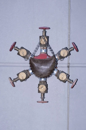 Brass industrial valve fixture with red accents hangs against a grey tiled wall, featuring six valves and chain supports.