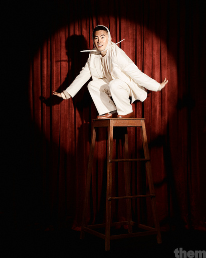Asian adult male in white suit crouching on wooden stool in stage spotlight with red curtain background in a theatrical pose.