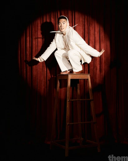 Asian adult male in white suit crouching on wooden stool in stage spotlight with red curtain background in a theatrical pose.