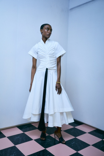 Elegant Black woman in a white layered dress poses in a studio with a pink and black tiled floor, showcasing a stylish and fashionable look.