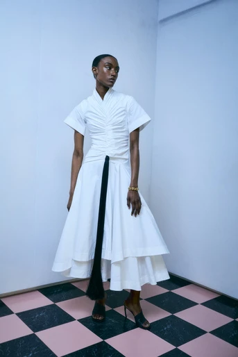 Elegant Black woman in a white layered dress poses in a studio with a pink and black tiled floor, showcasing a stylish and fashionable look.