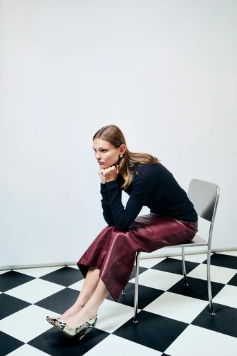 Adult woman in black sweater and wine-colored skirt sits on metal chair in studio with black-and-white checkered floor.