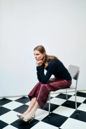 Adult woman in black sweater and wine-colored skirt sits on metal chair in studio with black-and-white checkered floor.