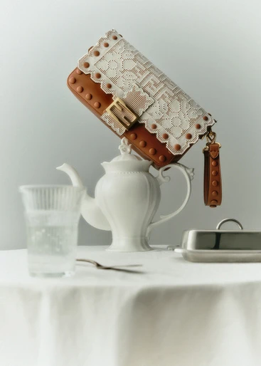 Designer handbag with lace detail balancing on a porcelain teapot beside a water glass and butter dish on a white tablecloth.