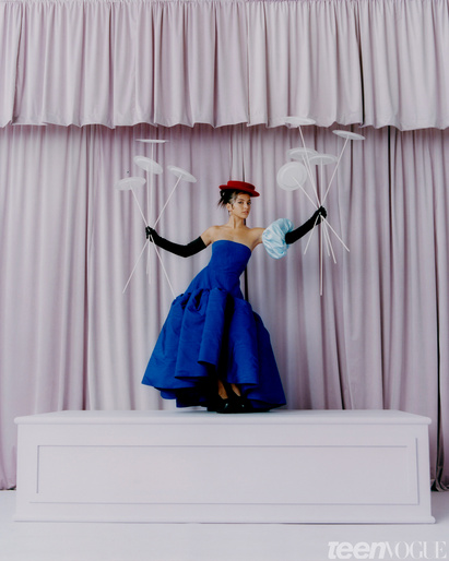 A model in a blue gown and black gloves poses on a platform in front of a pink curtain with paper disc props for a fashion shoot.