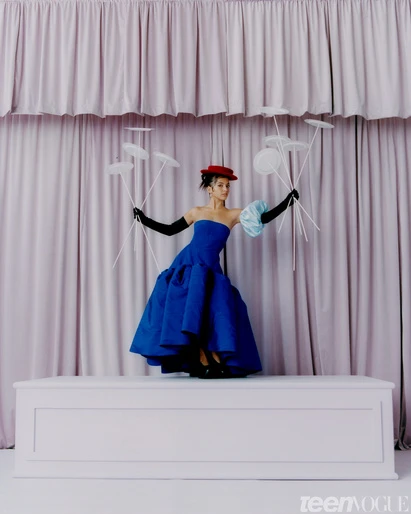 A model in a blue gown and black gloves poses on a platform in front of a pink curtain with paper disc props for a fashion shoot.