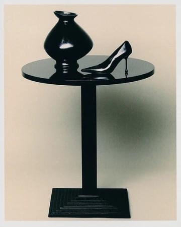 A black vase and a high-heeled shoe sit on a black table against a plain beige background, creating a minimalist still life.