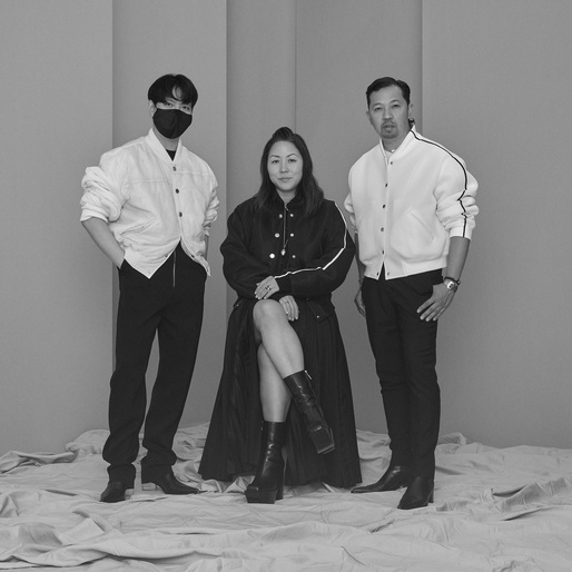 Black and white studio shot of three adults: one seated Asian woman in long coat, two standing Asian men in jackets and pants.