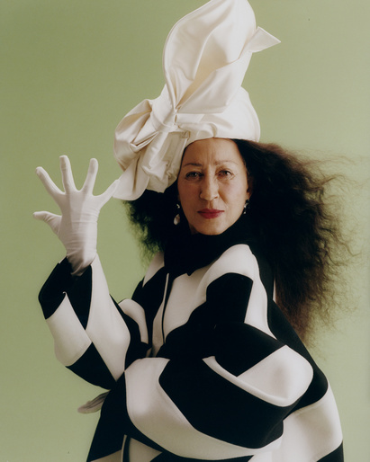 Elegant woman with dark, curly hair wears a black-and-white checker patterned jacket and large white hat against a light green backdrop.