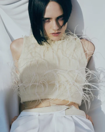 Model with dark bob hairstyle wearing a cream top embellished with white feathers against a white backdrop.