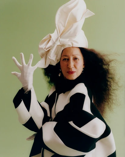 Woman wearing sculptural white hat and black and white patterned coat, raising a gloved hand against a green background.