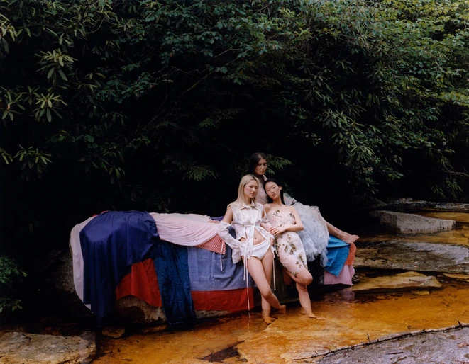 Three models posed by a fabric-draped rock in a stream surrounded by lush trees, set designed by Selena Liu.