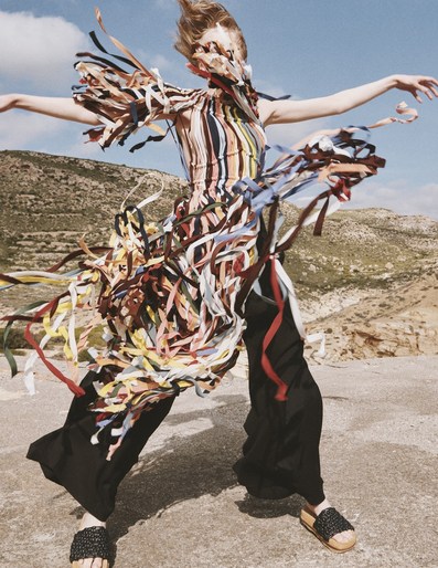 Fashionable adult female in black pants and ribbon-covered top strikes a pose against a background of hills on a bright, sunny day.