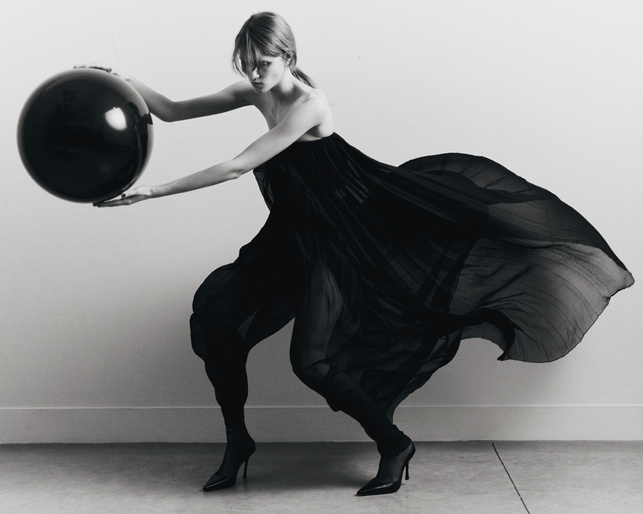 Black-and-white fashion shot: An adult female wearing a long, dark dress holds a large, spherical black ball in a minimalist studio setting.