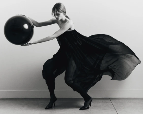 Black-and-white fashion shot: An adult female wearing a long, dark dress holds a large, spherical black ball in a minimalist studio setting.