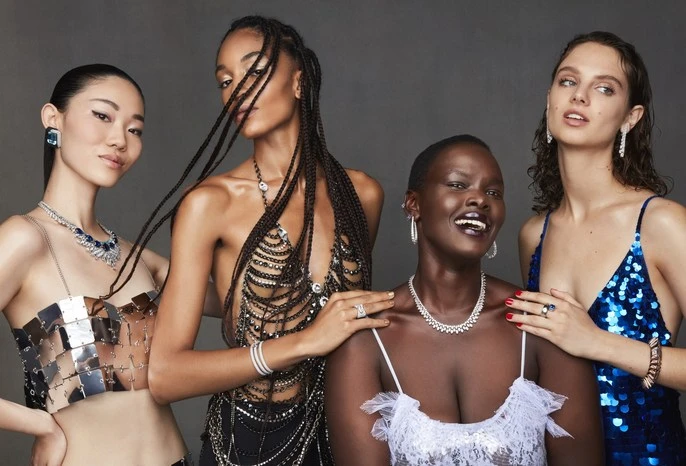 A group of four multi-ethnic adult women in a photo studio, wearing metallic and jeweled tops, posing for a fashion jewelry product shot.