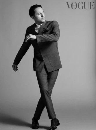 Monochrome portrait of a man in a tailored grey suit posing with crossed legs against a seamless studio backdrop.