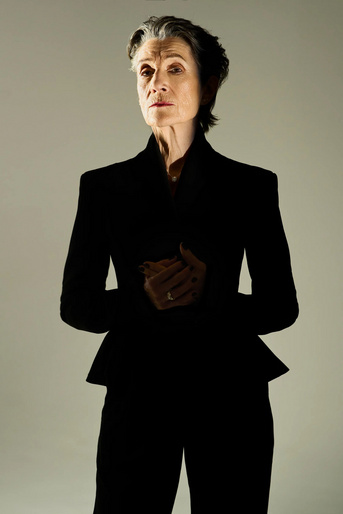 Elegant older woman in dark blazer and matching pants stands indoors with a neutral expression against a light grey backdrop.