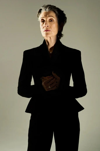 Elegant older woman in dark blazer and matching pants stands indoors with a neutral expression against a light grey backdrop.
