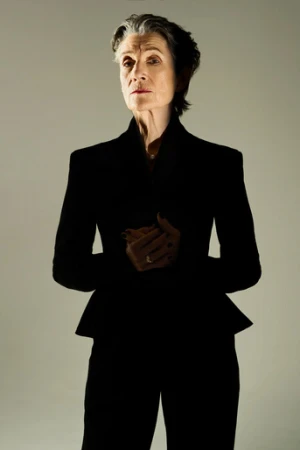 Elegant older woman in dark blazer and matching pants stands indoors with a neutral expression against a light grey backdrop.