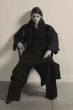 Model wearing a black oversized belted coat seated on a chair against a neutral background, styled by Julia Brenard.